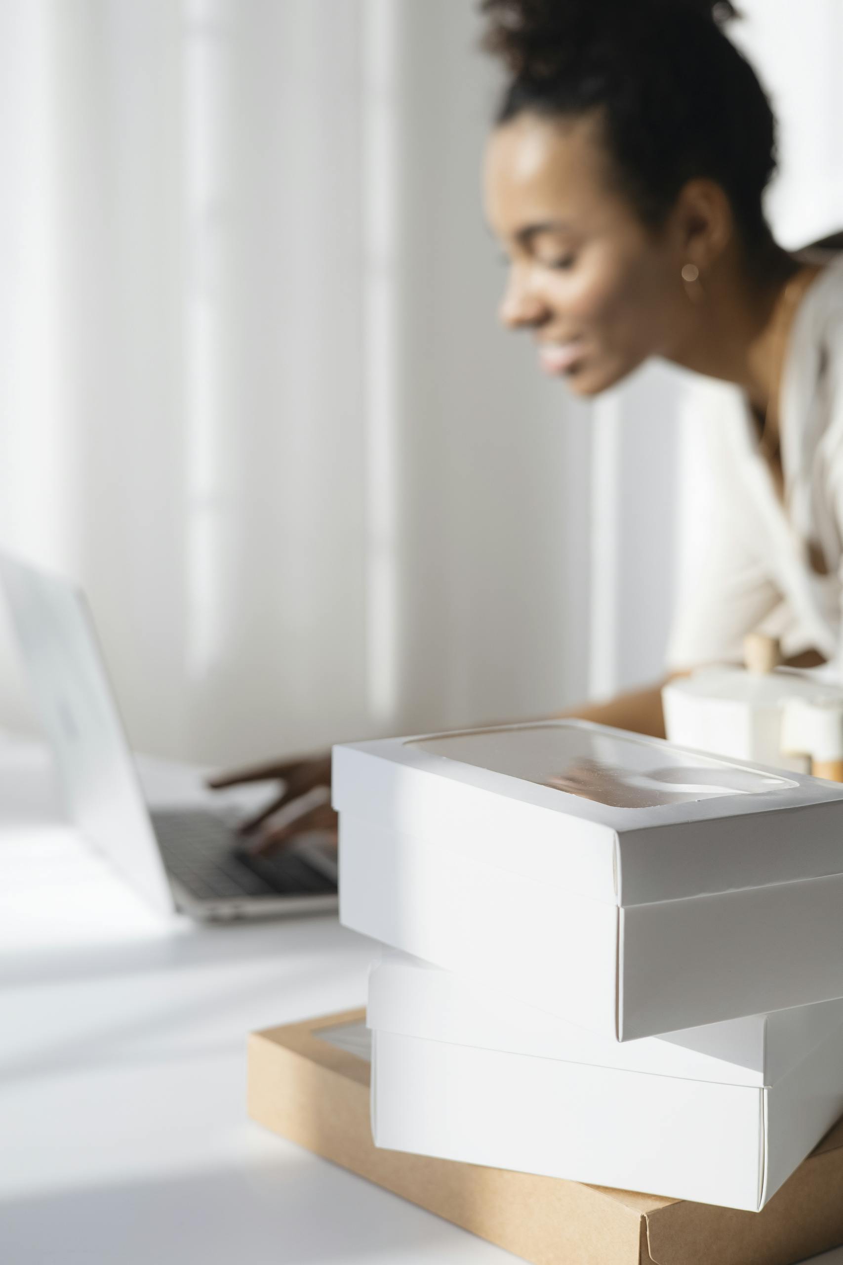 Eine jüngere Person of Color mit heller Bluse sitze vor hellem Hintergrund vor einem weißen Notebook. im Vordergrund unscharf die Ecken von Boxen