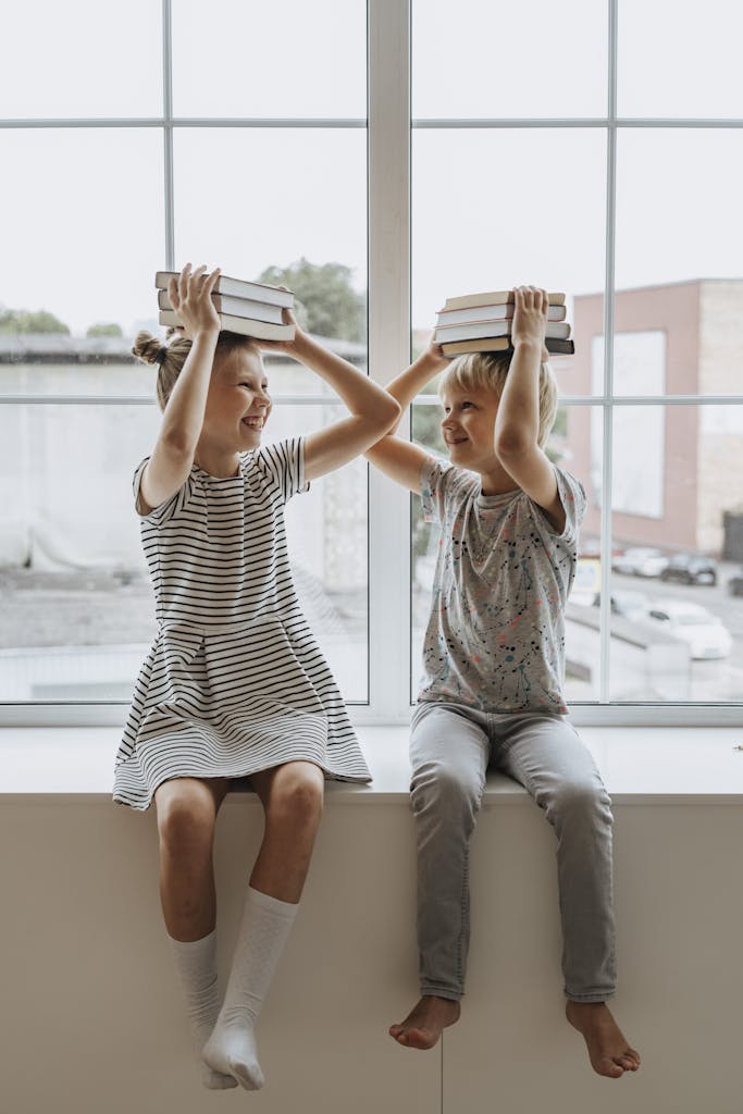 Zwei Grundschul-Kinder in heller Kleidung sitzen auf einem Fensterbrett vor einer breiten und hellen Fensterfront und jonglieren Bücher auf ihrem Kopf.