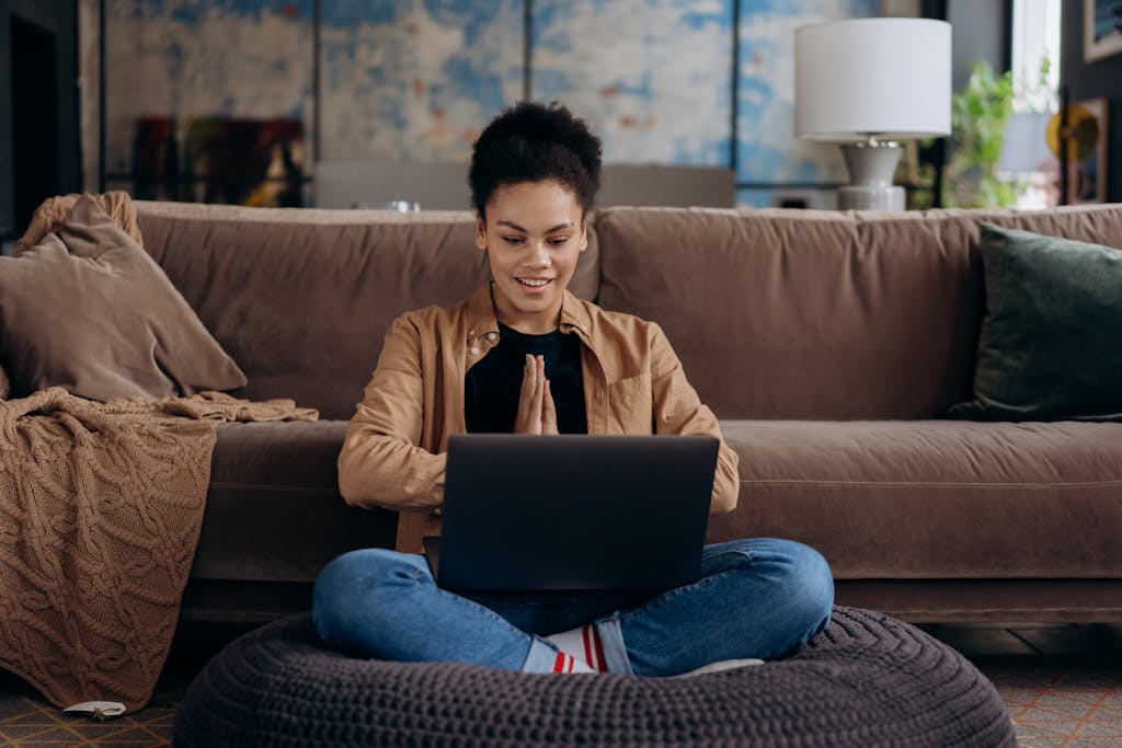 Eine lächelnde junge Frau sitzt im Schneidersitz auf einem Sofa und benutzt ihren Laptop in einer gemütlichen Wohnumgebung.